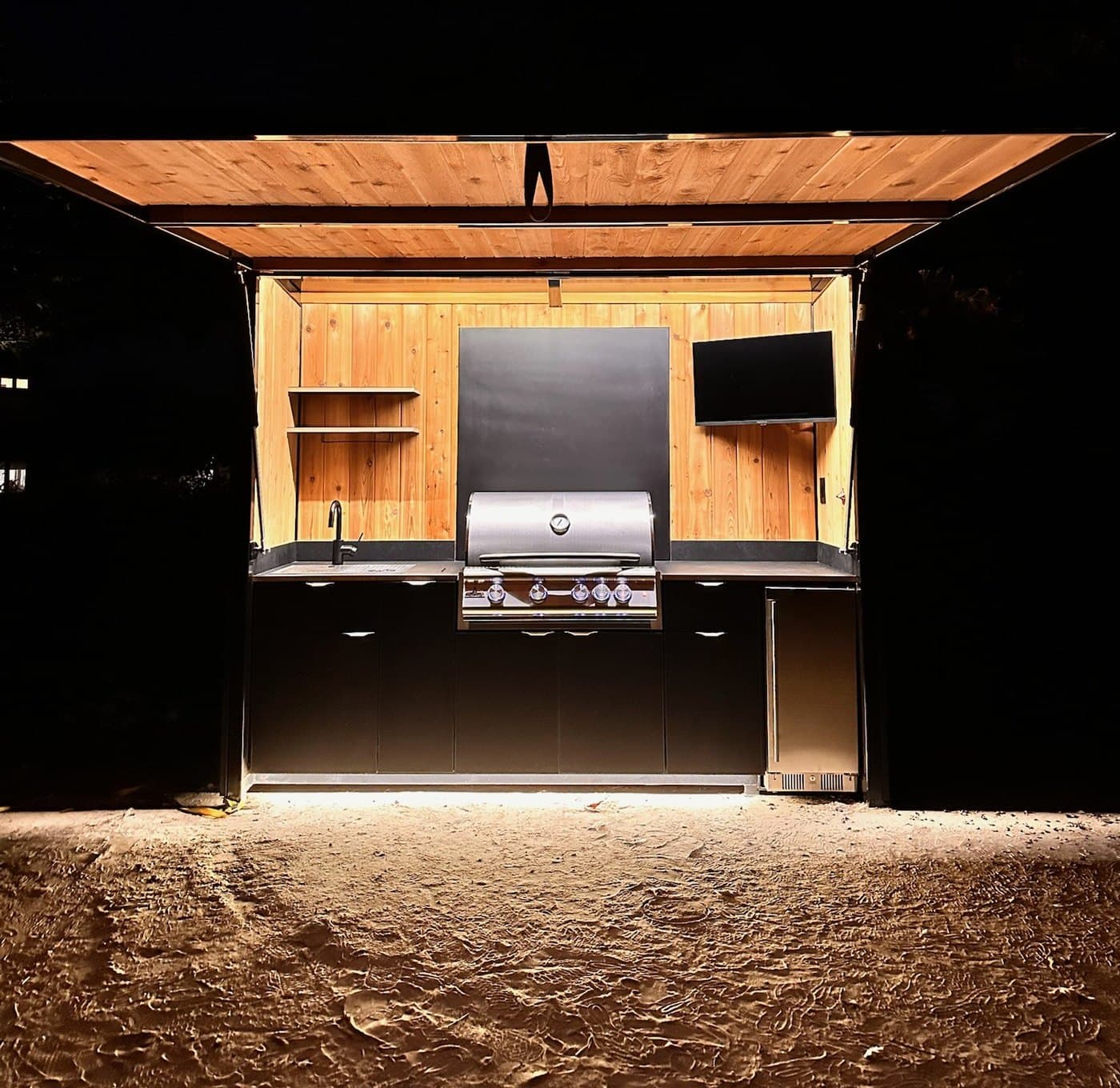 Enclosed outdoor kitchen pod open and lit at night, awning raised, grill and interior aglow